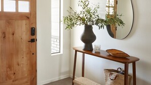 A wooden door, a console table with a vase and mirror, a woven basket, and a patterned rug.