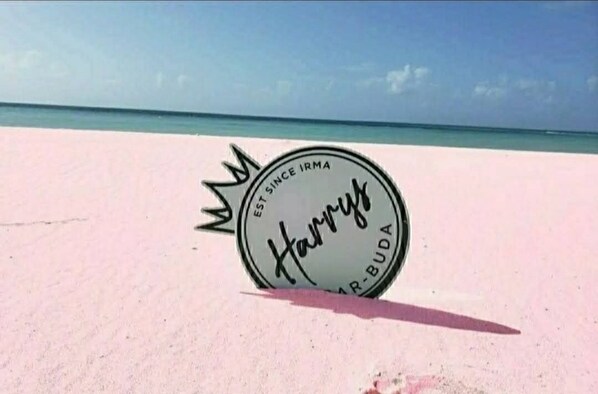 A circular sign with the text "Harry's Bar-Buda" on a sandy beach with a clear blue sky and ocean in the background.