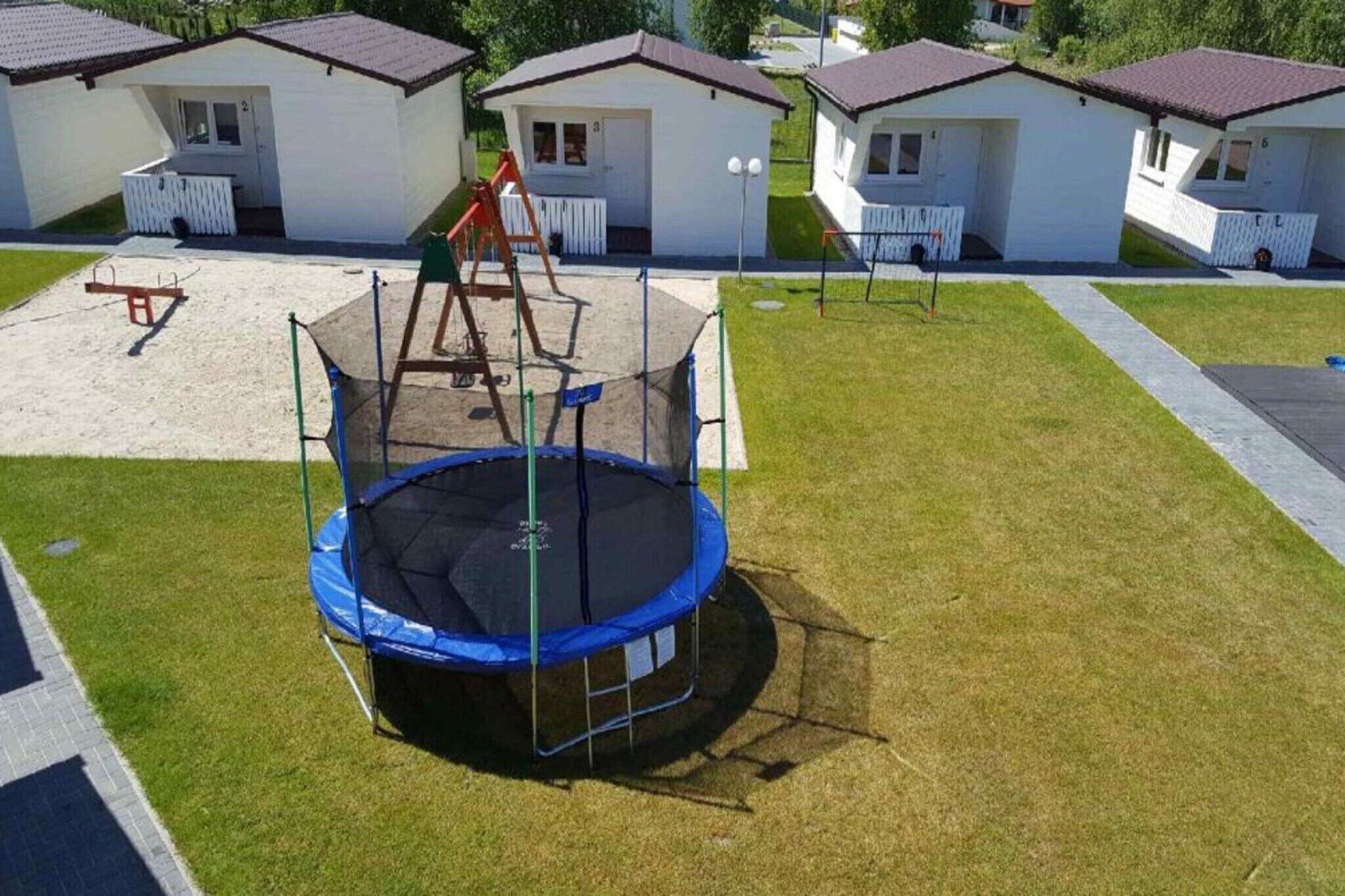 A playground with a trampoline and swings, surrounded by small white cabins with brown roofs.
