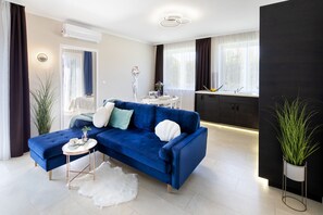 Un soggiorno moderno con un divano blu, un tappeto bianco e morbido, un piccolo tavolino rotondo e un angolo cottura con lavello e pensili.