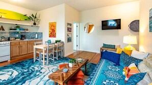 A modern living room with a sofa, coffee table, and TV. There is a kitchen area with wooden cabinets and a surfboard.