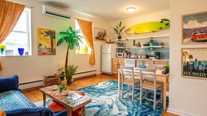 A cozy living room with a sofa, a coffee table, and a dining area with a table and chairs. There is a kitchen area with a microwave, stove, and refrigerator. A surfboard is displayed on the wall, and there are various decorative items and plants.
