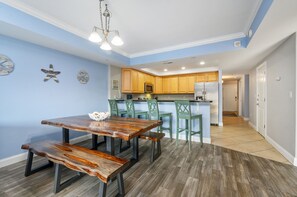 A modern kitchen with wooden cabinets, a central island, and a dining area with a wooden table and benches. - Sunset Beach 208 by Seaside Vacations & Sales (Ocean City)
