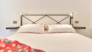 Una cama bien hecha con ropa de cama blanca y detalles rojos, un cabecero con luces de lectura integradas y mesitas de noche con lámparas.