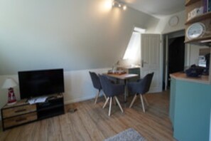 A compact living space with a television, a dining table with chairs, a small kitchenette, and a clock on the wall. - Ferienwohnung in Wyk-Boldixum auf Foehr (Wyk-Boldixum)