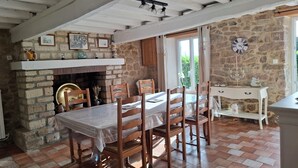 Un comedor con chimenea de ladrillo, sillas de madera, una mesa con mantel de encaje y pared de piedra con un reloj.