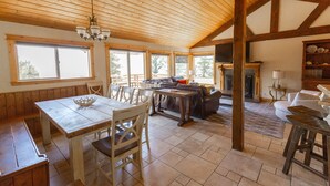 Amplia sala de estar con techo de madera, ventanas grandes, chimenea y una mesa de comedor con bancos.