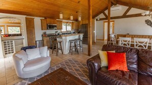 Amplia sala de estar con un sofá de cuero, un sillón blanco y una mesa de comedor con sillas. Se ve una cocina con gabinetes de madera y una isla central al fondo.
