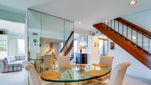 Un espace repas moderne avec une table en verre et quatre chaises, un escalier en bois, et un salon avec un canapé et une table à manger en arrière-plan.