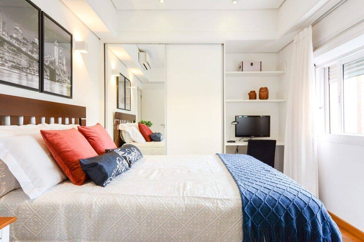 Quarto de hotel com cama, escrivaninha com computador, televisão e estante de livros.