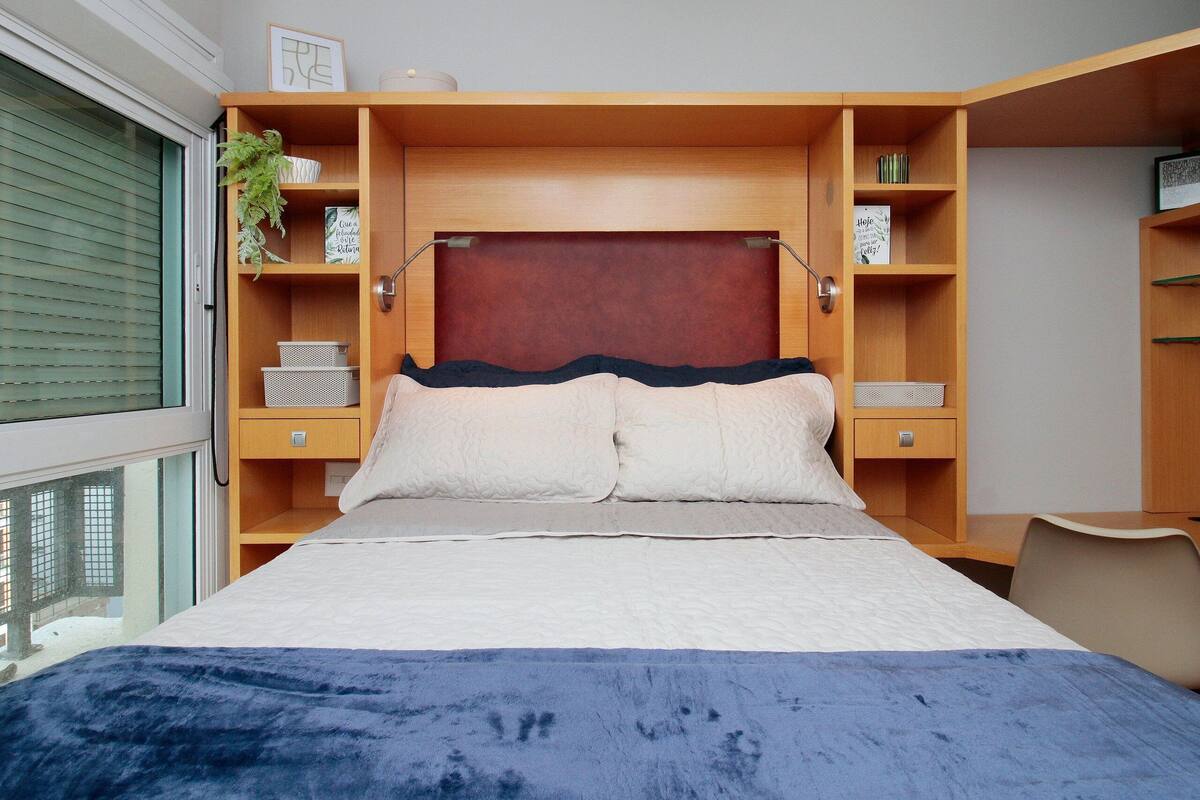 Quarto com cama grande, cabeceira de madeira, prateleiras embutidas e janela com persianas.
