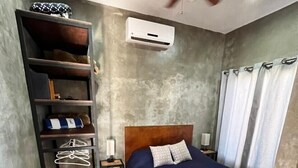 Een slaapkamer met een bed, een plafondventilator, een plank met spullen, een stoel en een luchtverkoeler aan de muur.