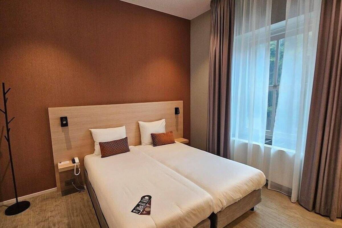 Ein Hotelzimmer mit einem großen Bett, einem Nachttisch, einer Lampe, einem Fenster mit Vorhängen und einem Wandbild.