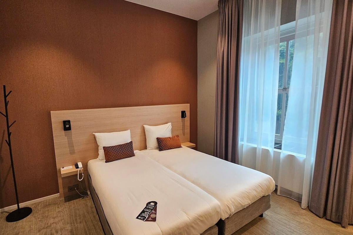 Ein Hotelzimmer mit einem großen Bett, einem Nachttisch, einer Lampe und einem Fenster mit Vorhängen.