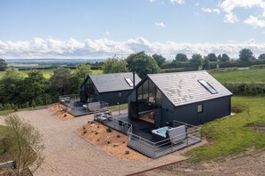 A modern two-story house with a large deck and a hot tub, surrounded by a rural landscape.