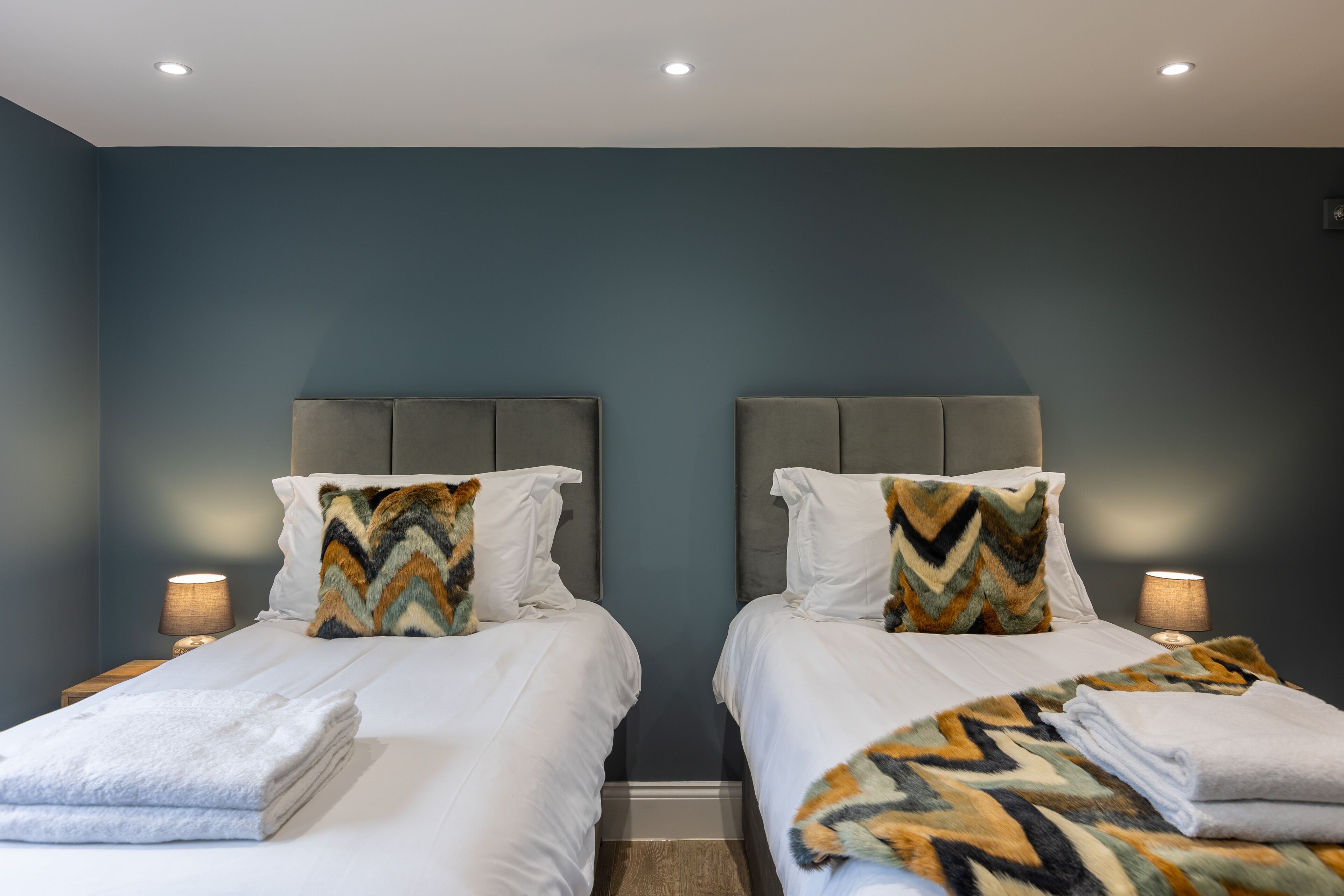A hotel room with two beds, each with a grey headboard, white bedding, and a patterned throw blanket. There are bedside lamps and a small table with a lamp.