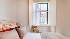 A hotel room with a bed, pillows, a chair, a window with curtains, and a view of a building outside. - 1BR Apartment - Downtown Lisbon - Chiado (Lisbon)