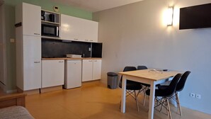Une cuisine compacte avec des meubles blancs, un four à micro-ondes et un petit réfrigérateur. On y trouve une table à manger avec quatre chaises et une poubelle.