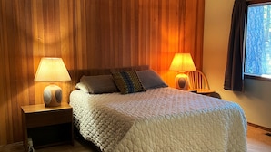 A bedroom with a large bed, two bedside tables with lamps, wooden paneling on the walls, and a window with curtains. - RR18 National Forest Views! (Black Butte Ranch)