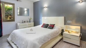 Une chambre moderne avec un grand lit, des tables de chevet, un miroir et une fenêtre donnant sur un paysage verdoyant.
