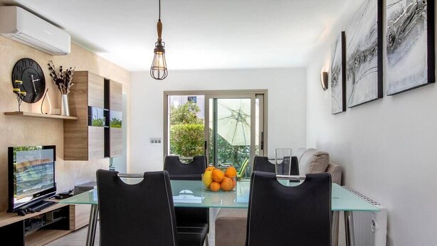 Un salon moderne avec une table à manger en verre, des chaises noires, un téléviseur et une fenêtre donnant sur un paysage verdoyant.