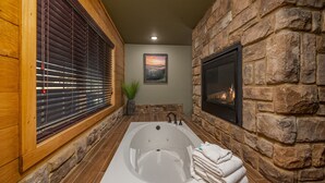 A bathroom with a stone wall, a large bathtub, wooden blinds, a fireplace, and a framed picture on the wall. - Heaps O'fun (Gatlinburg)