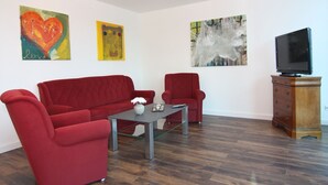 Een woonkamer met een rode bank, een rode fauteuil, een salontafel en een tv op een houten kast. Er hangen drie schilderijen aan de muur.