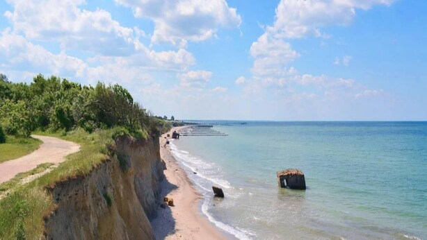 Une route côtière menant à une plage, avec une falaise et un grand rocher dans l’eau.