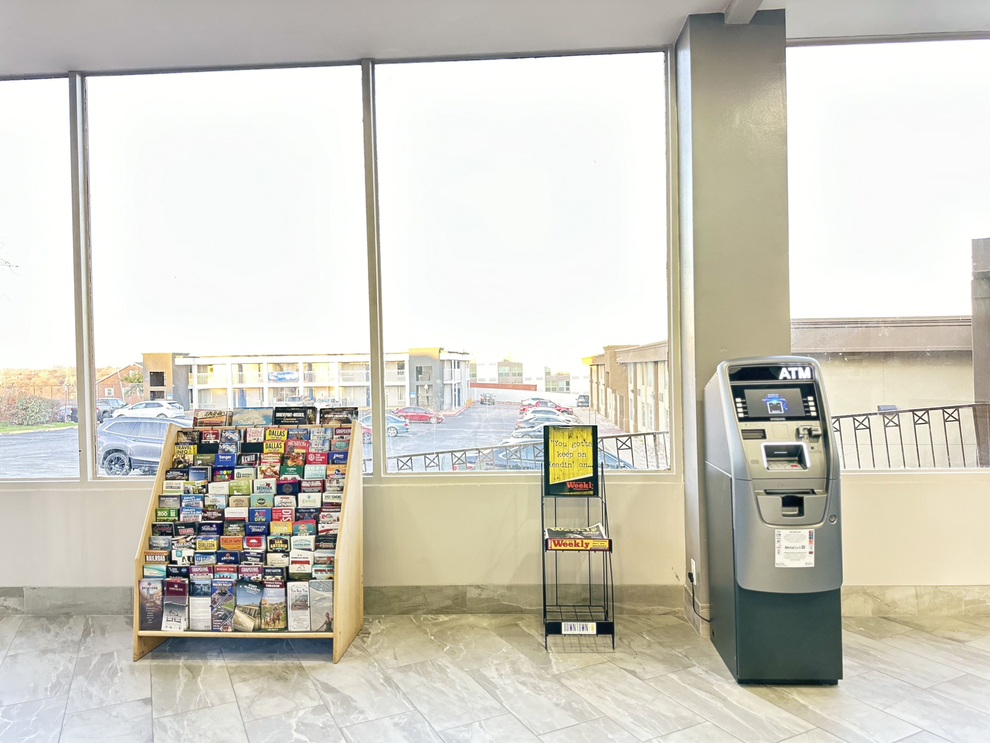 A window with a view of a parking lot and a building, a magazine rack, and an ATM machine inside a building.