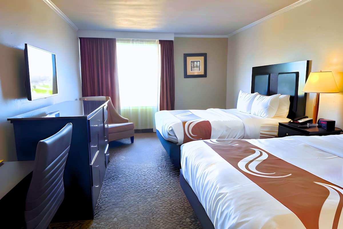 A hotel room with a desk, two beds, a chair, a TV, a lamp, and a framed picture on the wall.