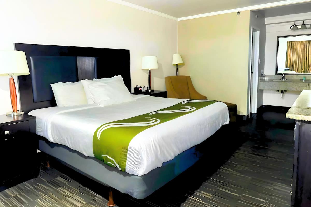 A hotel room with a large bed, two bedside tables, a chair, a desk, a mirror, and a bathroom with a sink and mirror.