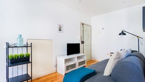Un salon moderne avec un canapé gris, un meuble de rangement blanc pour appareils électroniques, une étagère en métal noir ornée de plantes et un lampadaire noir.