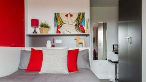 Un dormitorio moderno con una cama gris, almohadas rojas y grises, un plaid rojo, una lámpara roja, una planta, una escultura de un caballo de madera y un cuadro en la pared.