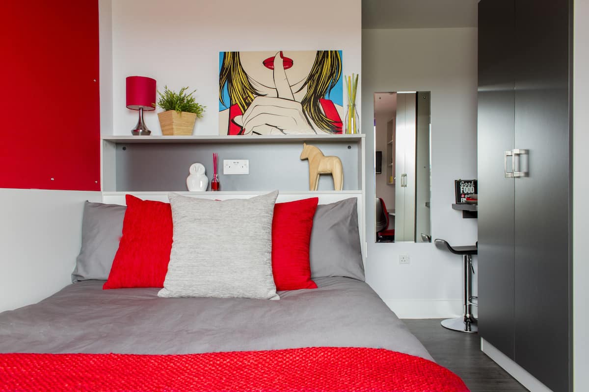 Quarto moderno com cama cinza, almofadas vermelhas e cinzas, manta vermelha, luminária vermelha, uma planta, escultura de um cavalo de madeira e um quadro na parede.