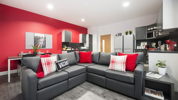 Una sala moderna con un sofá seccional grisáceo, cojines rojos, un comedor con una mesa blanca y una cocina con armarios grises y electrodomésticos blancos.