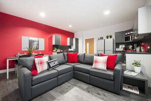 Una sala moderna con un sofá seccional grisáceo, cojines rojos, un comedor con una mesa blanca y una cocina con armarios grises y electrodomésticos blancos.