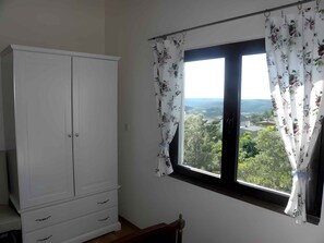 Une pièce avec une armoire blanche, une fenêtre ornée de rideaux à motifs floraux et une vue sur la verdure et les bâtiments.