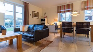 Una sala de estar moderna con un sofá, una mesa de comedor y una silla. Hay una ventana con cortinas, una lámpara y una estantería.