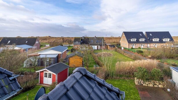 Un barrio residencial con varias casas, algunas con techos inclinados y otras con techos planos, rodeado de campos verdes y un cielo despejado.