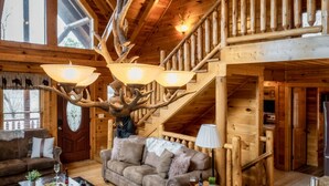 A log cabin interior with a large antler chandelier, a staircase, and a cozy living area with a couch and fireplace. - NERF War-Family Lodge-Theater-HotTub-GlowParty (Sevierville)