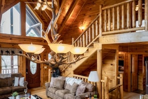 A log cabin interior with a large antler chandelier, a staircase, and a cozy living area with a couch and fireplace.