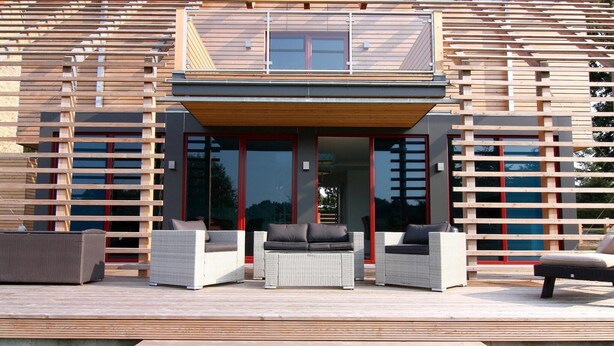 Una casa moderna con terraza de madera, mobiliario exterior y ventanas grandes.