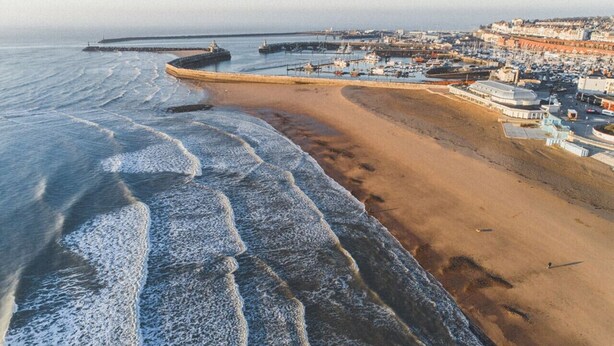 Une ville côtière dotée d’une plage de sable, d’une jetée et d’un port de plaisance où sont amarrés des bateaux.