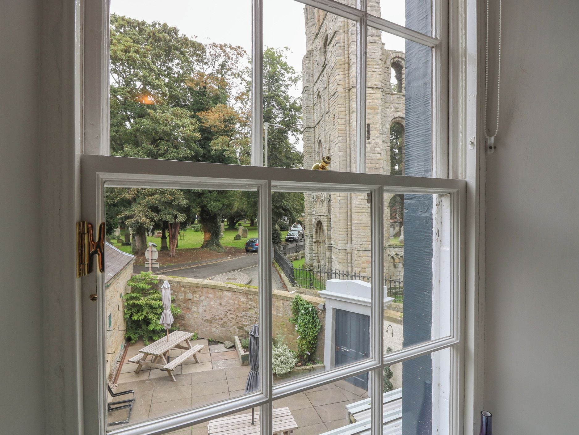 A view through a window of a courtyard with stone walls, a bench, and a building with arched windows.