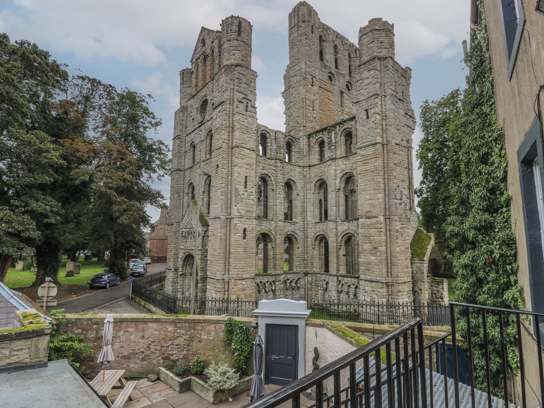 A historic stone building with multiple towers and arched windows, surrounded by trees and a paved area with a bench.