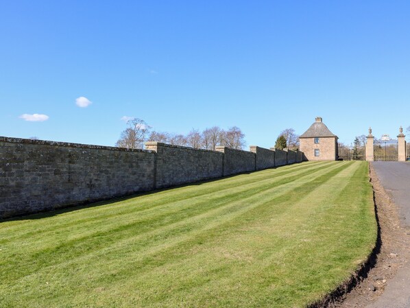 A stone wall with a grassy area in front, a building with a pitched roof, and a gated entrance.
