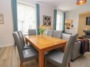 A dining area with a wooden table and grey chairs, a vase with flowers, and a sofa in the background.