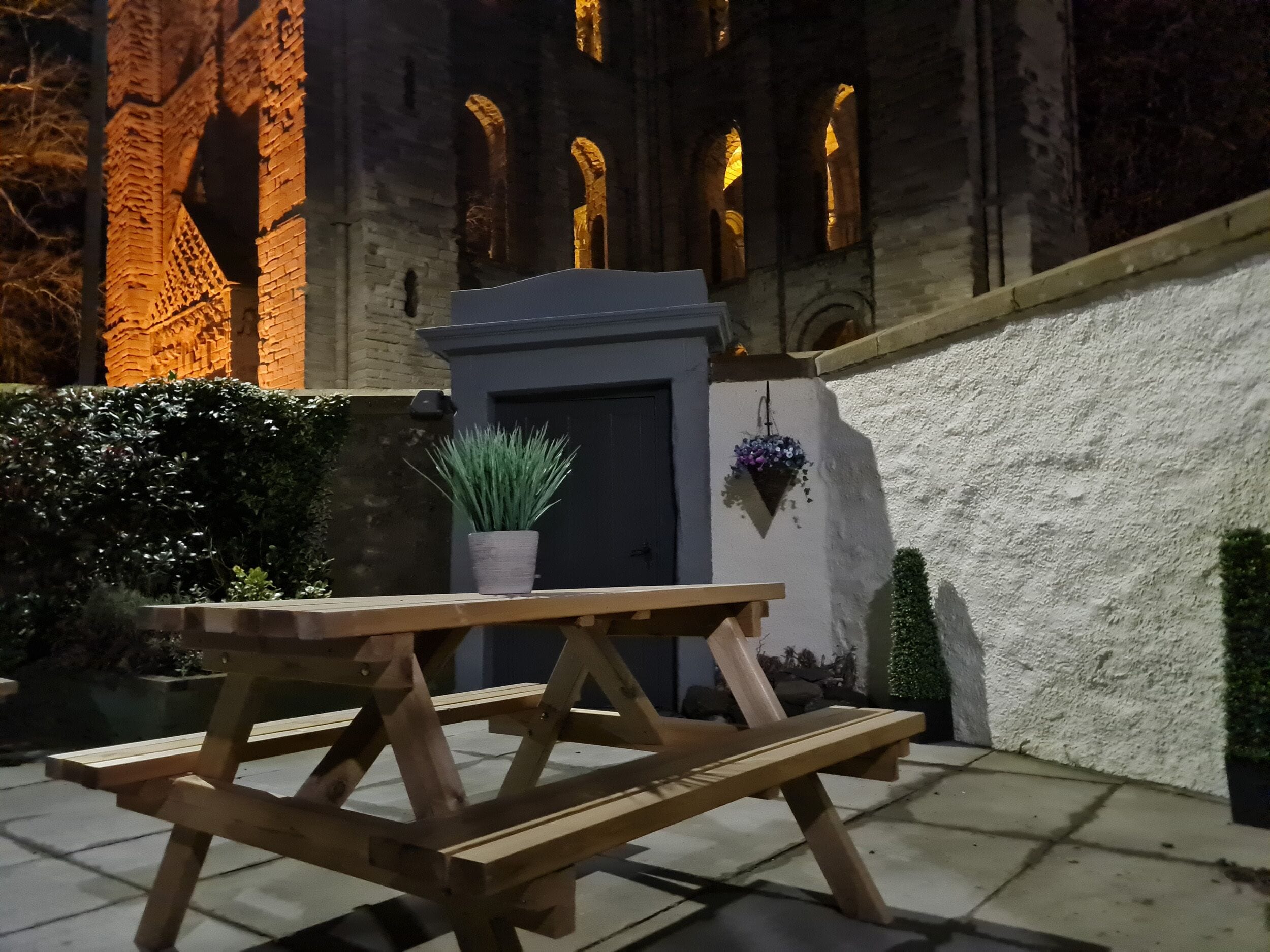 A wooden picnic table with a potted plant in front of a building with arched windows and a door.