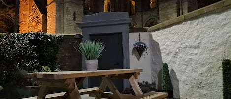 Uma mesa de piquenique de madeira com uma planta em vaso, em frente a um edifício com janelas e porta em arco.
