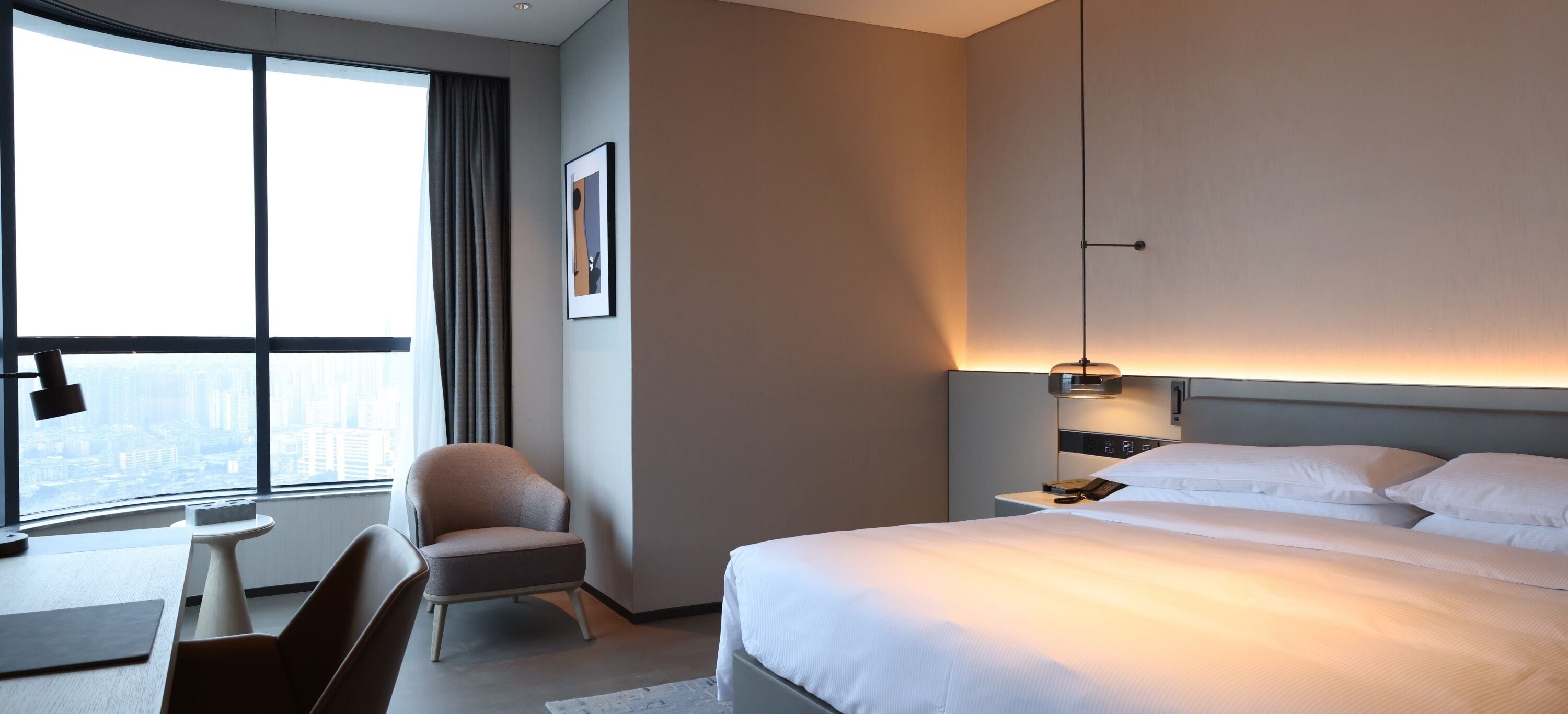 A modern hotel room with a large bed, a desk, a chair, and a view of the city through large windows.